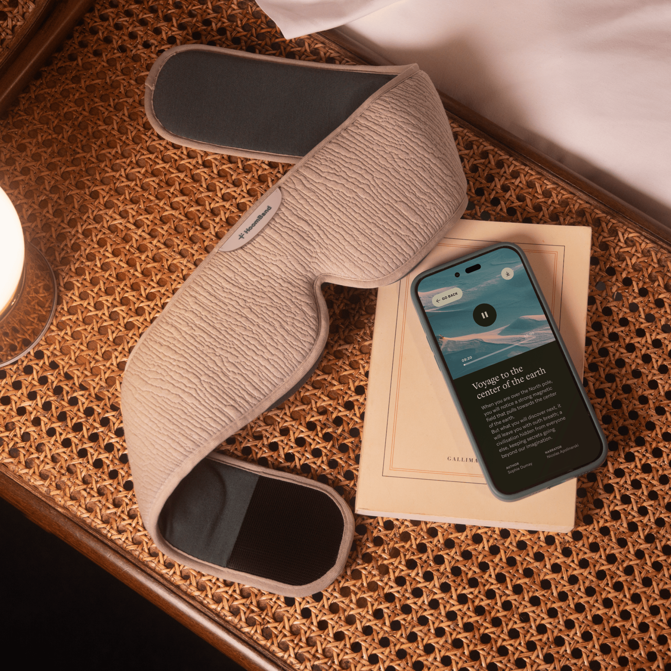 LIVLAB HoomBand Ultimate audio sleep mask with Bluetooth, resting on a nightstand beside a lamp and book.