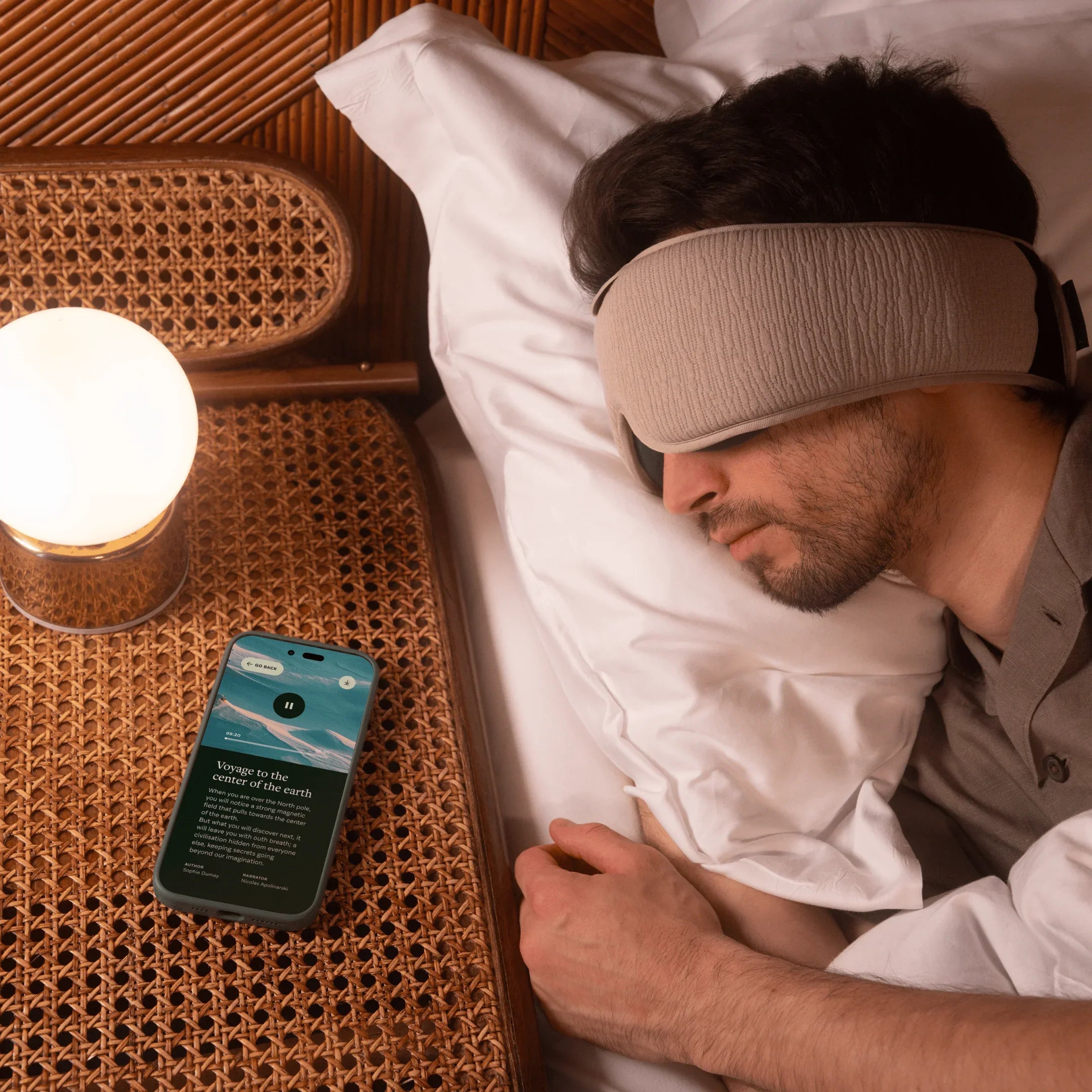Man sleeping on bed with sleep mask and audio headband, smartphone on nightstand playing sleep story, warm lamp light