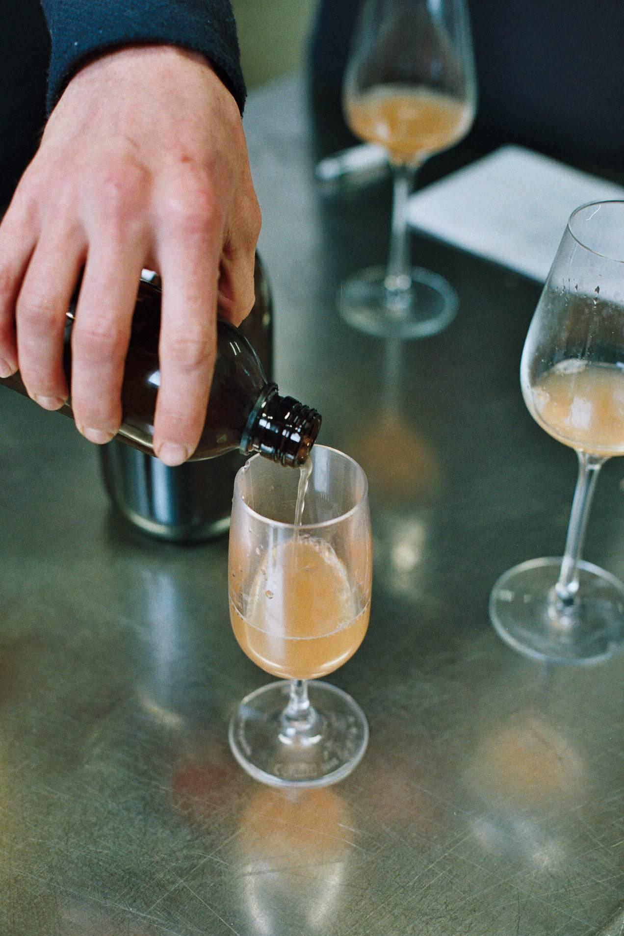 Hand pouring orange beverage from bottle into wine glass on metal table