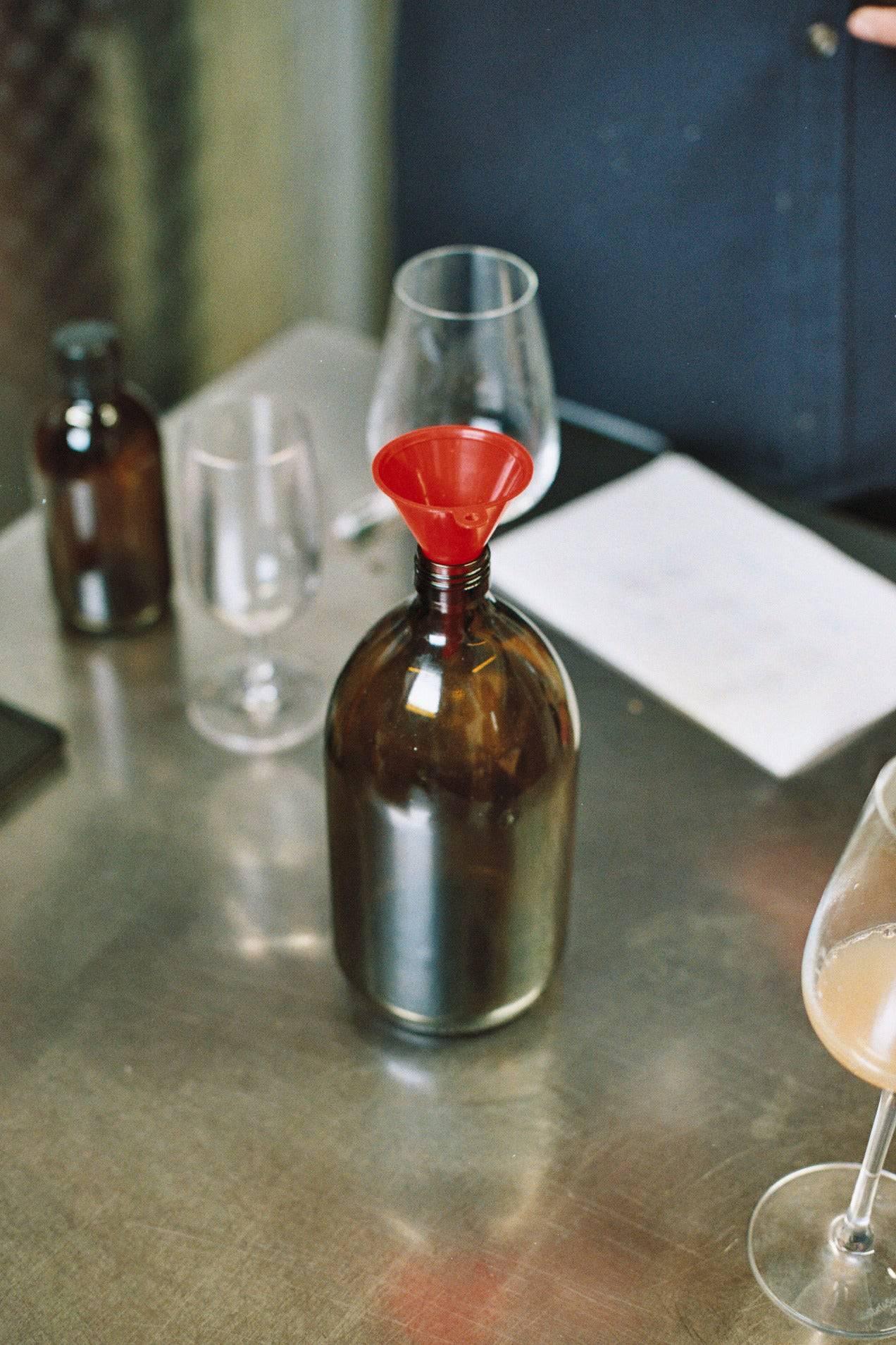 Brown glass bottle with red funnel on metal table, surrounded by empty glasses